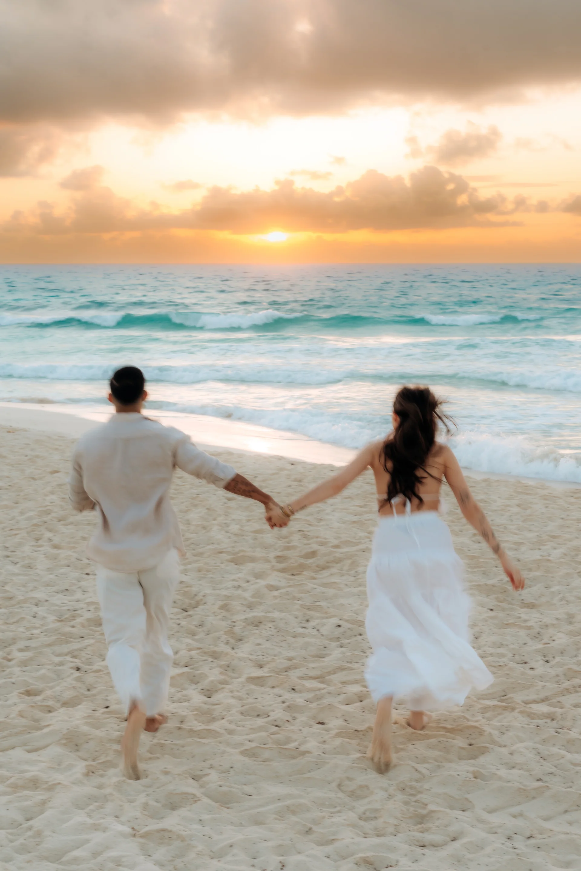 Couple seen from behind running hand in hand toward the sunrise over the sea, Playa Delfines, Cancún