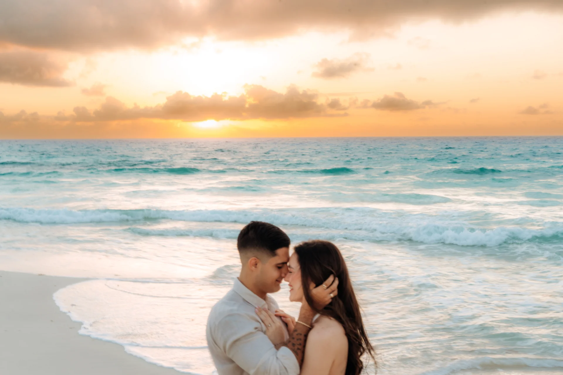 Couple standing forehead to forehead at the water's edge under a glowing sunrise sky, Playa Delfines, Cancún