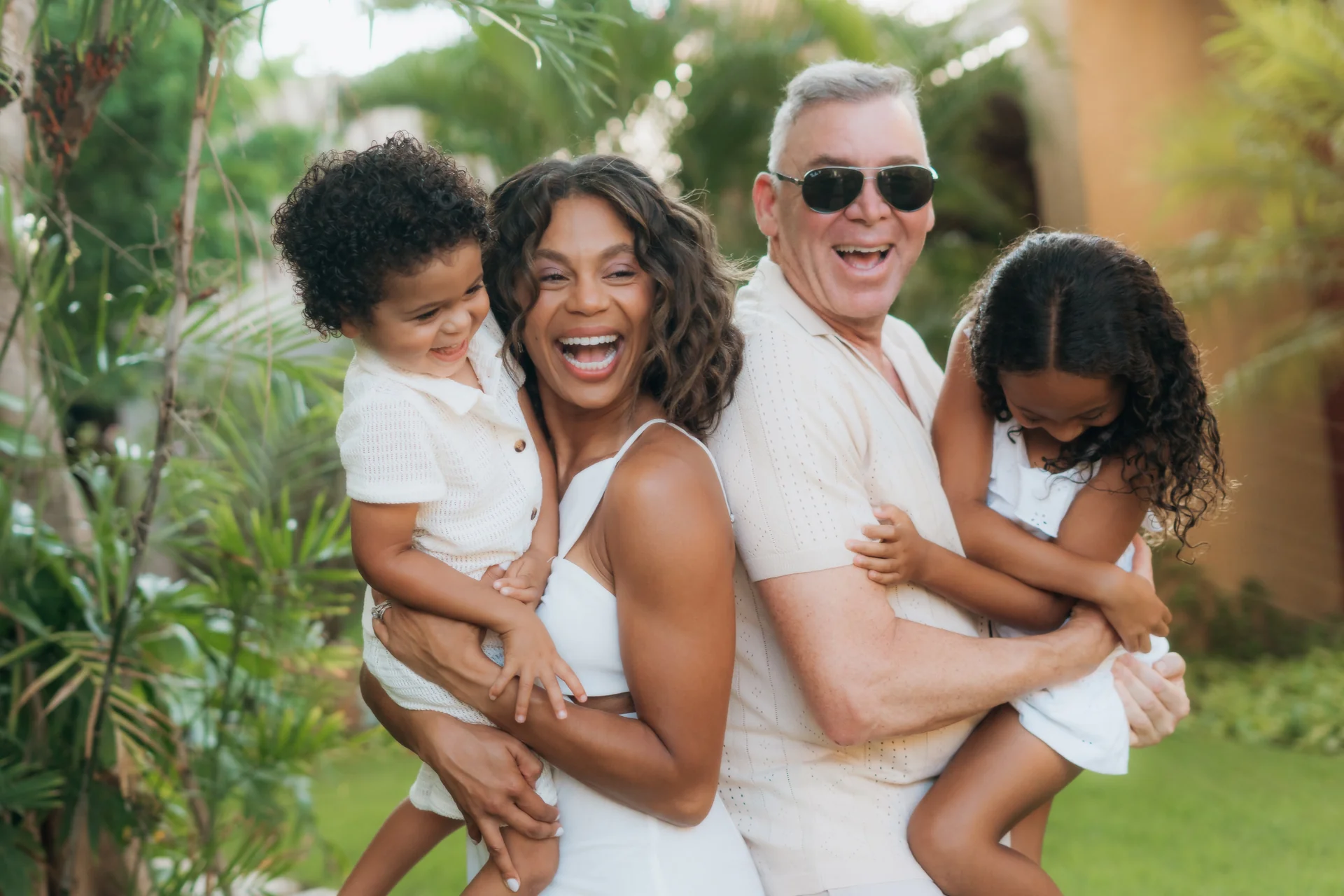 Laughing family of four in white, each parent carrying a child, among tropical plants, Banyan Tree Mayakoba, Riviera Maya