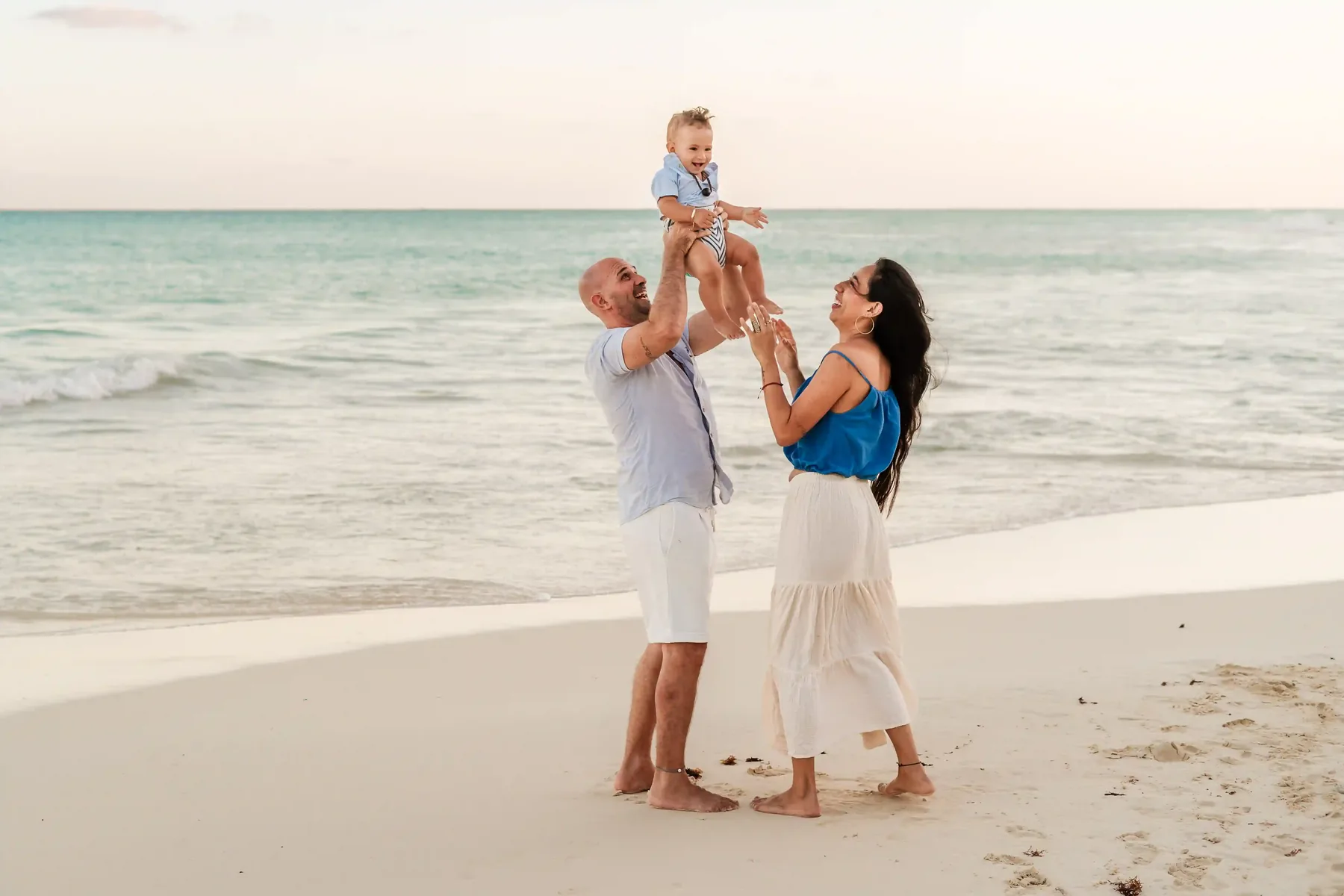 Parents holding baby on Cancún beach during family photoshoot at golden hour, mother laughing