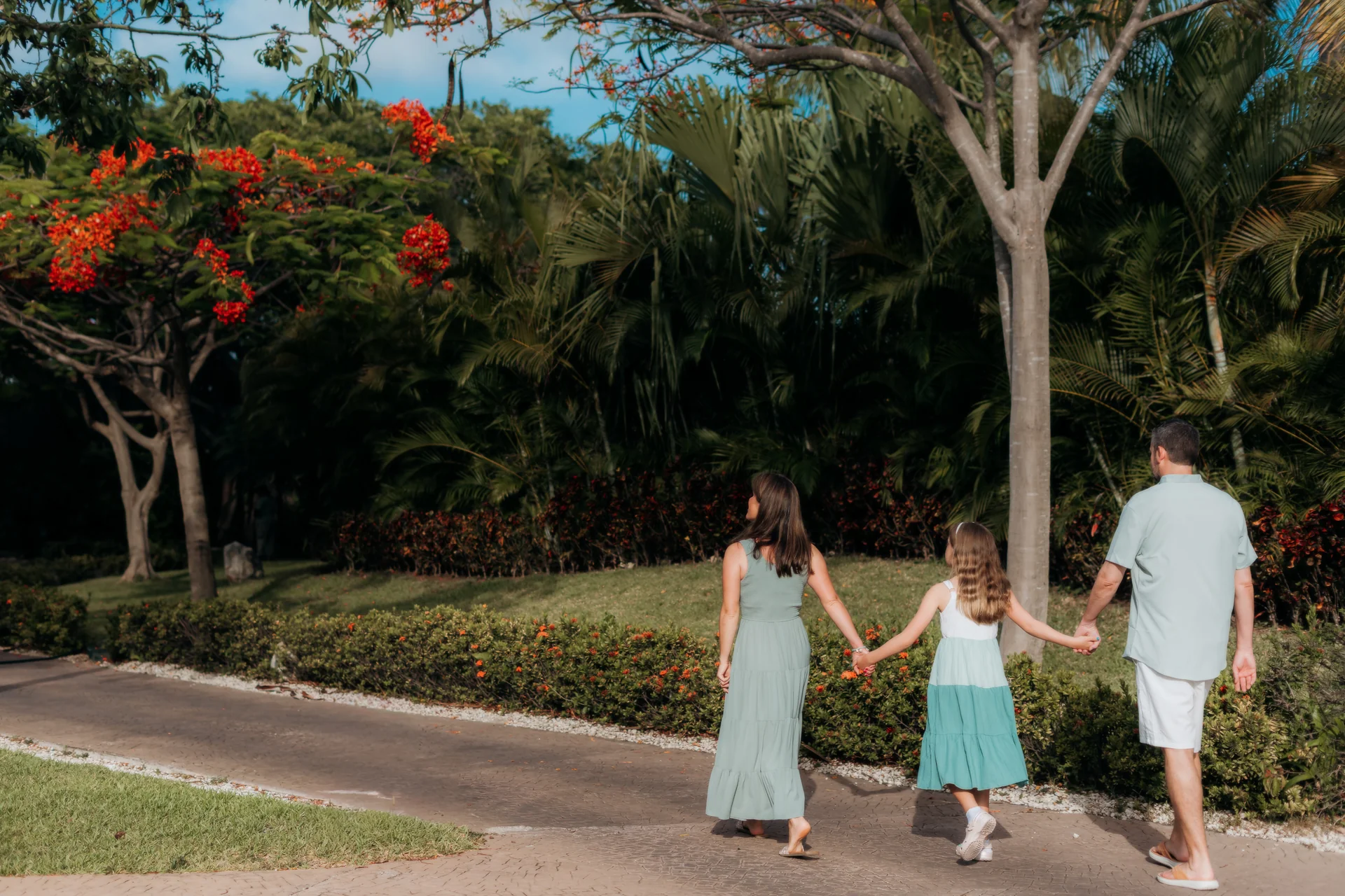 Family of three seen from behind holding hands on a path past flame trees and palms, Rosewood Mayakoba, Riviera Maya