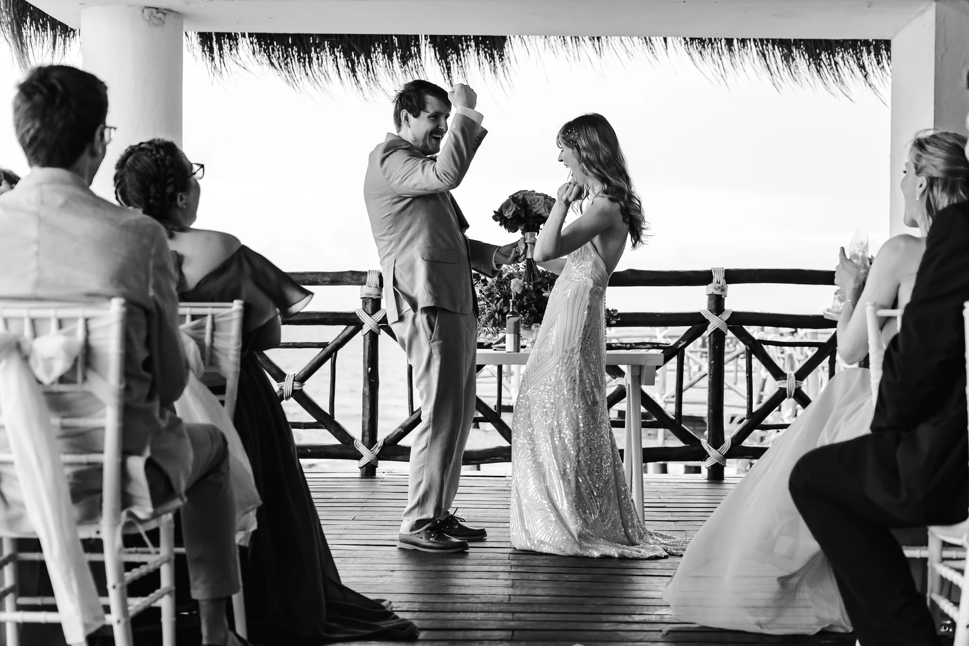 Black and white photograph of a bride and groom celebrating with raised fists at the ceremony table while seated guests watch