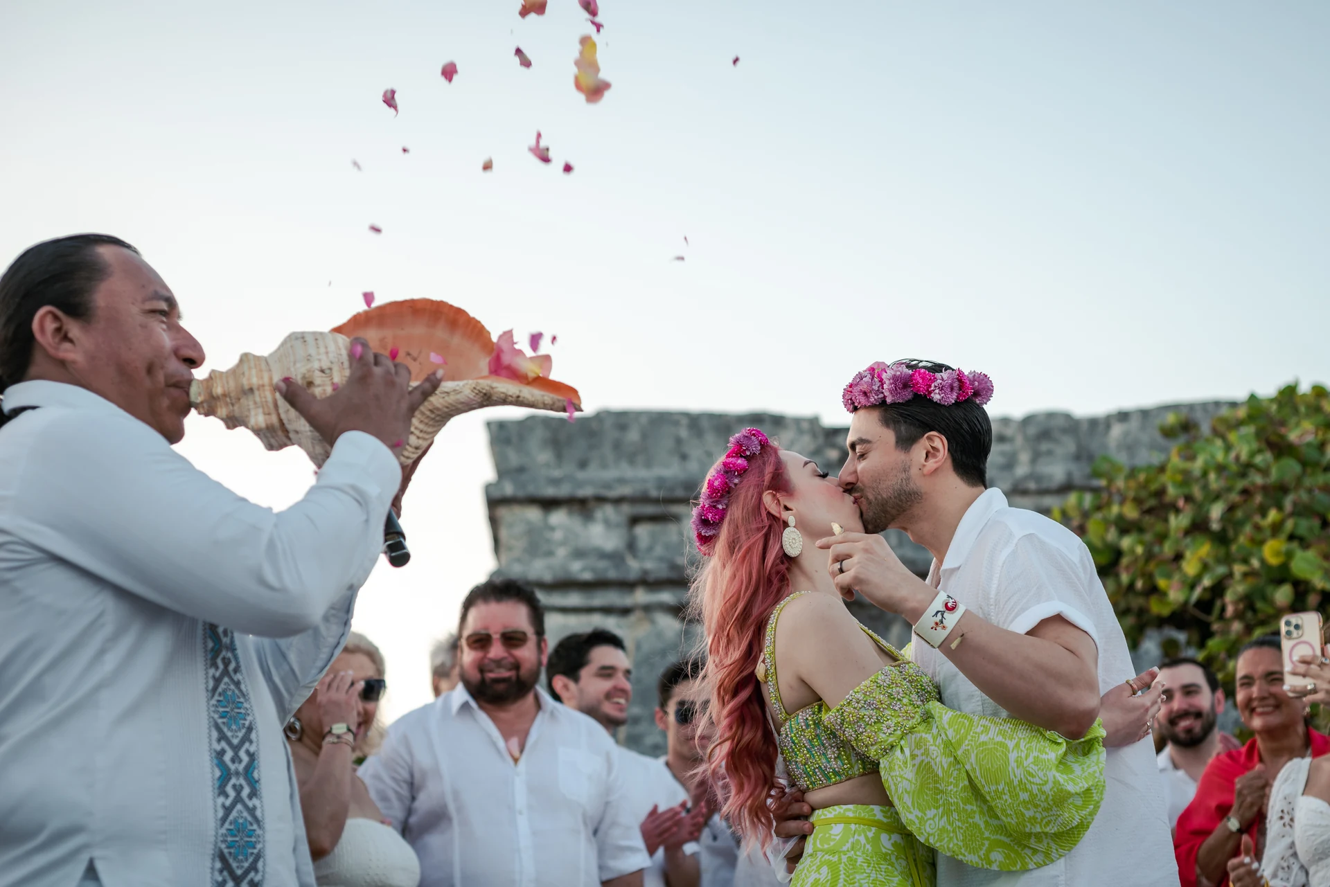 Bride and groom kissing in flower crowns while an officiant blows a conch shell under falling petals