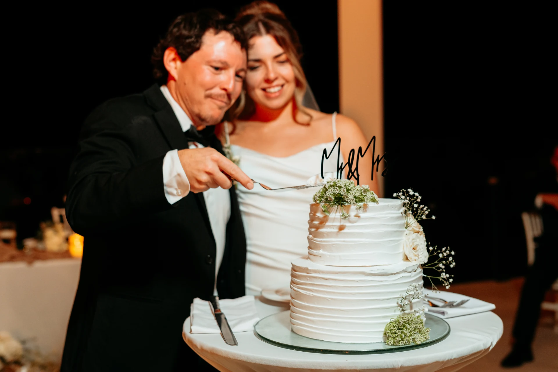 Bride and groom smiling as they cut their two-tier wedding cake together at night