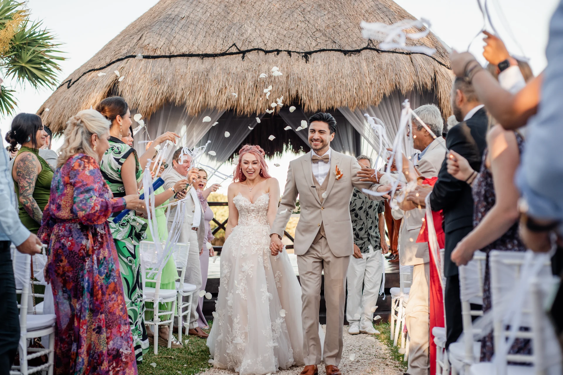 Bride and groom walking down the aisle under a palapa while guests throw white streamers and petals