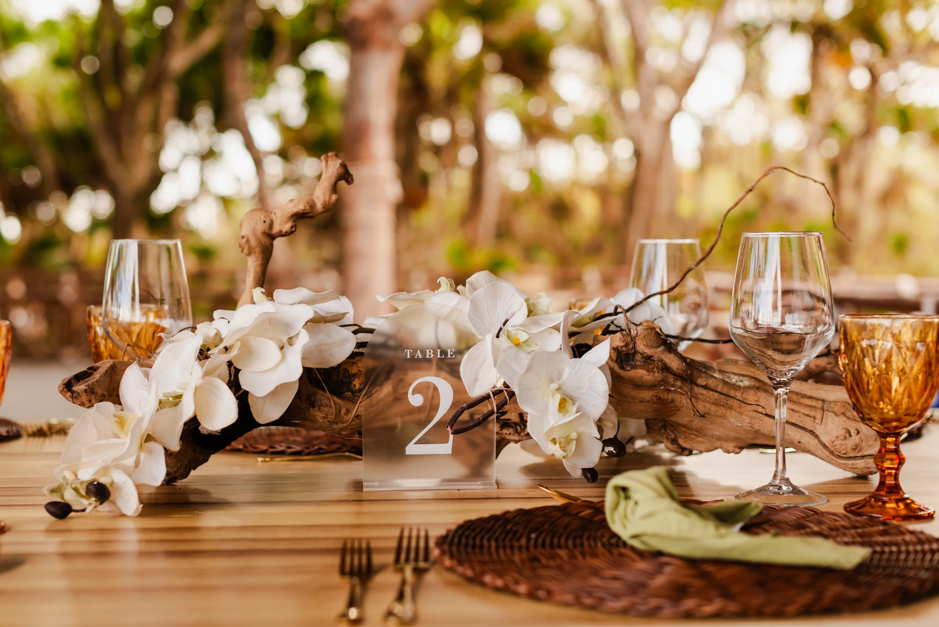 Wooden reception table centerpiece with white orchids, driftwood, wine glasses, and amber goblets