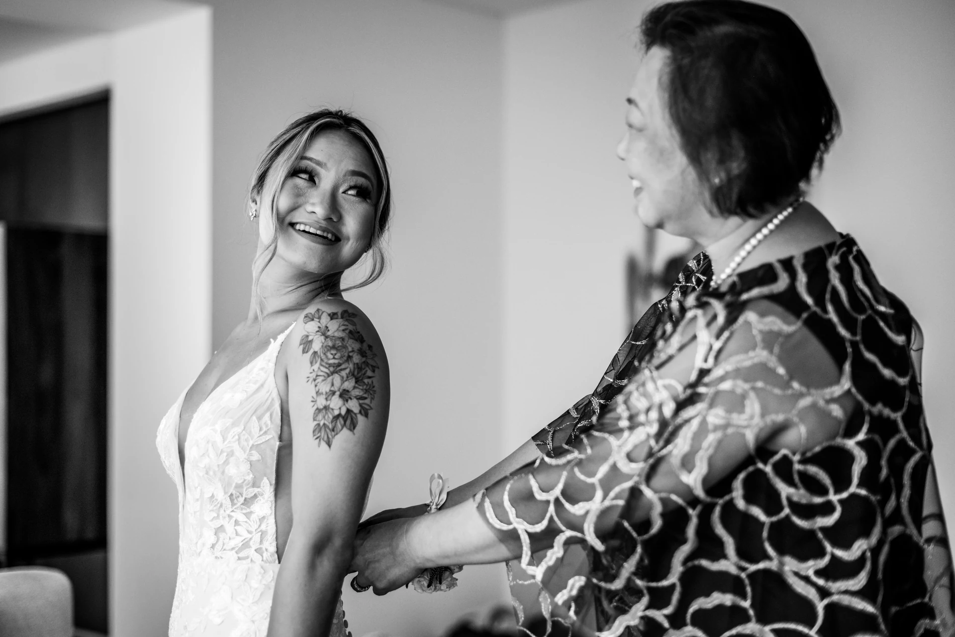 Bride smiling over her shoulder while her mother fastens the back of her wedding dress