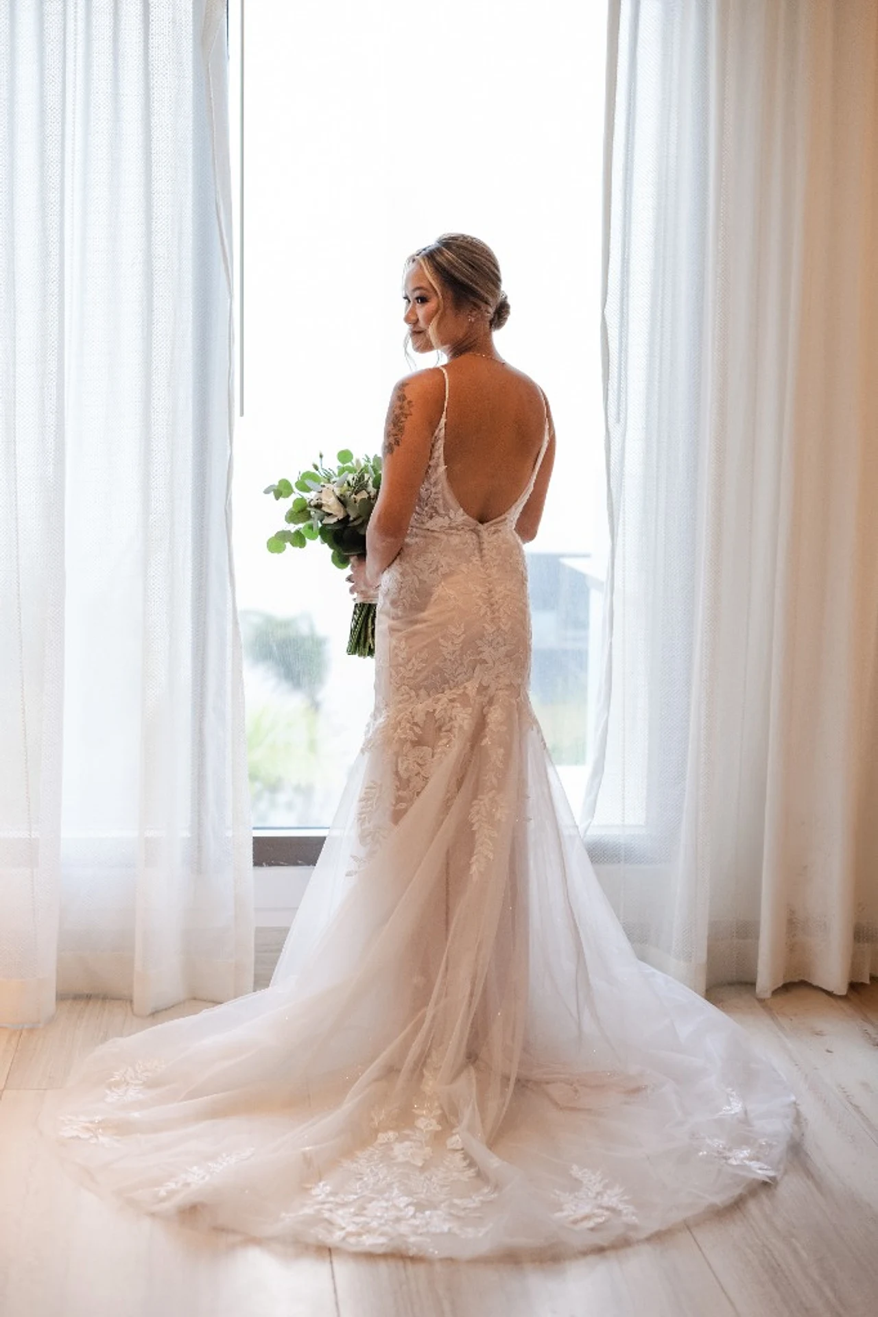 Bride in an open-back lace dress holding her bouquet in front of sheer window curtains