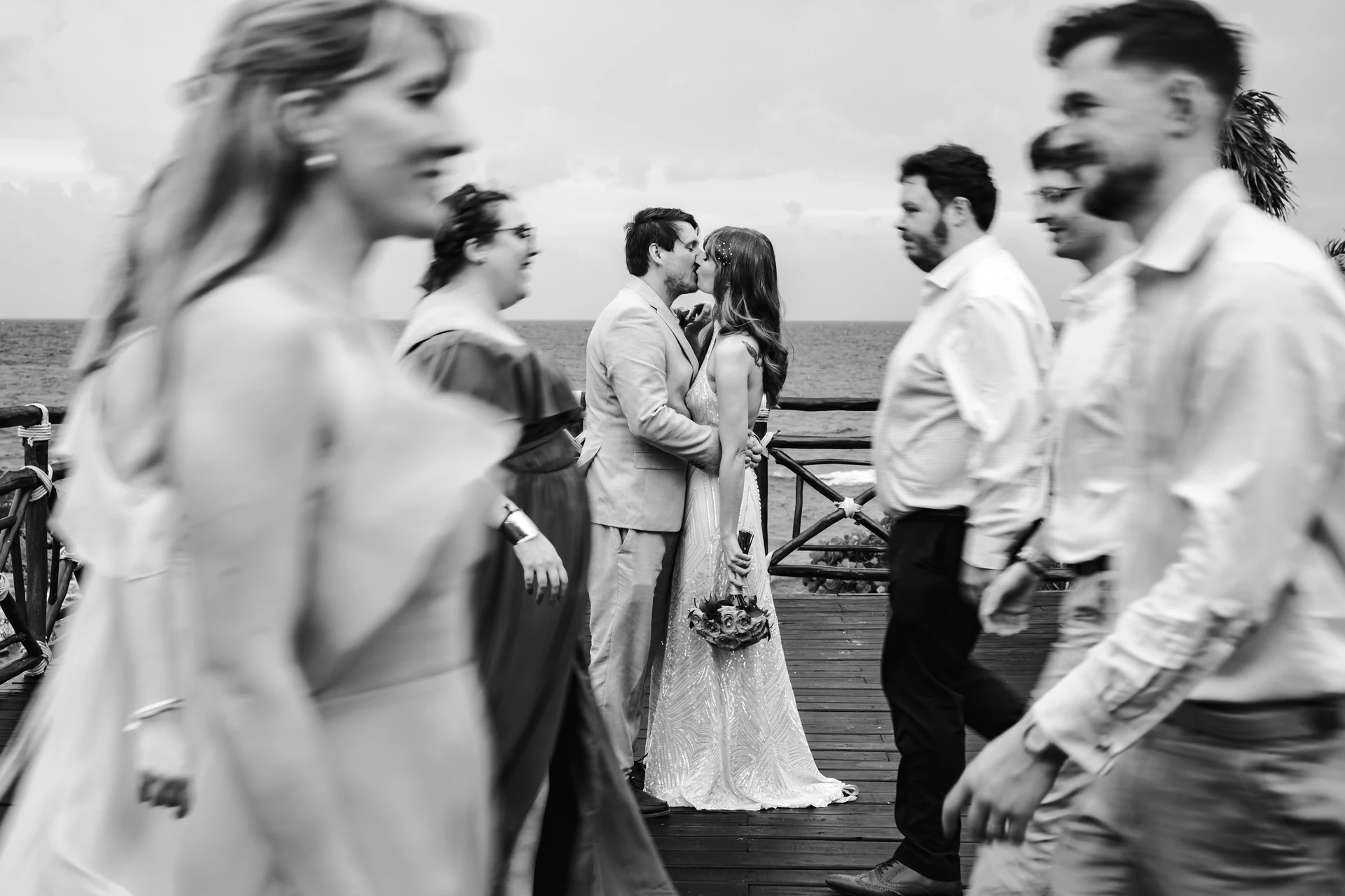 Fotografía en blanco y negro de una pareja que se besa en un muelle de madera frente al mar, mientras los invitados pasan en movimiento