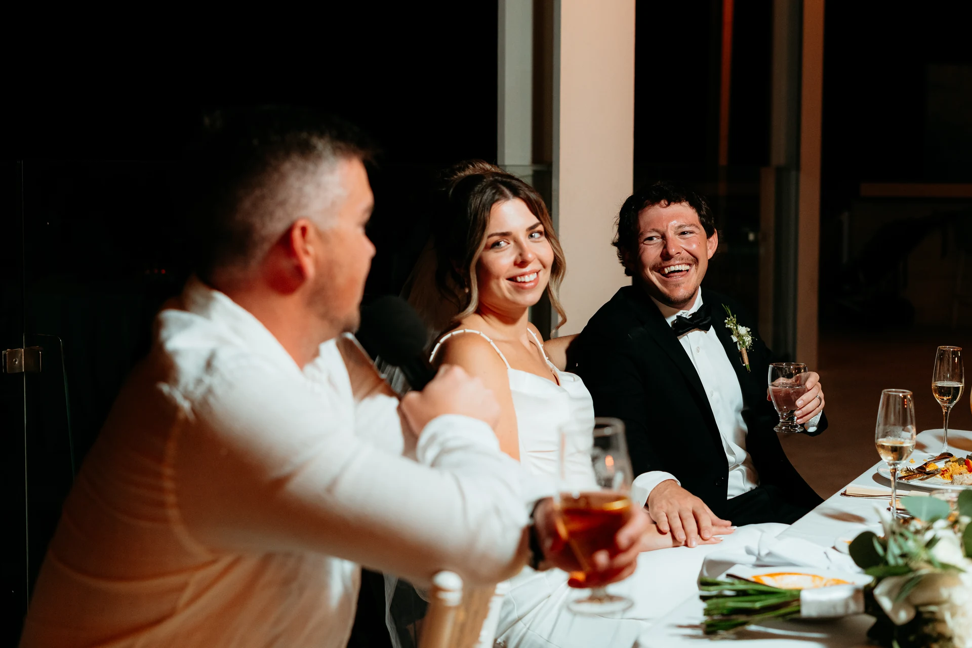Bride and groom laughing at the reception table while a guest gives a speech with a microphone