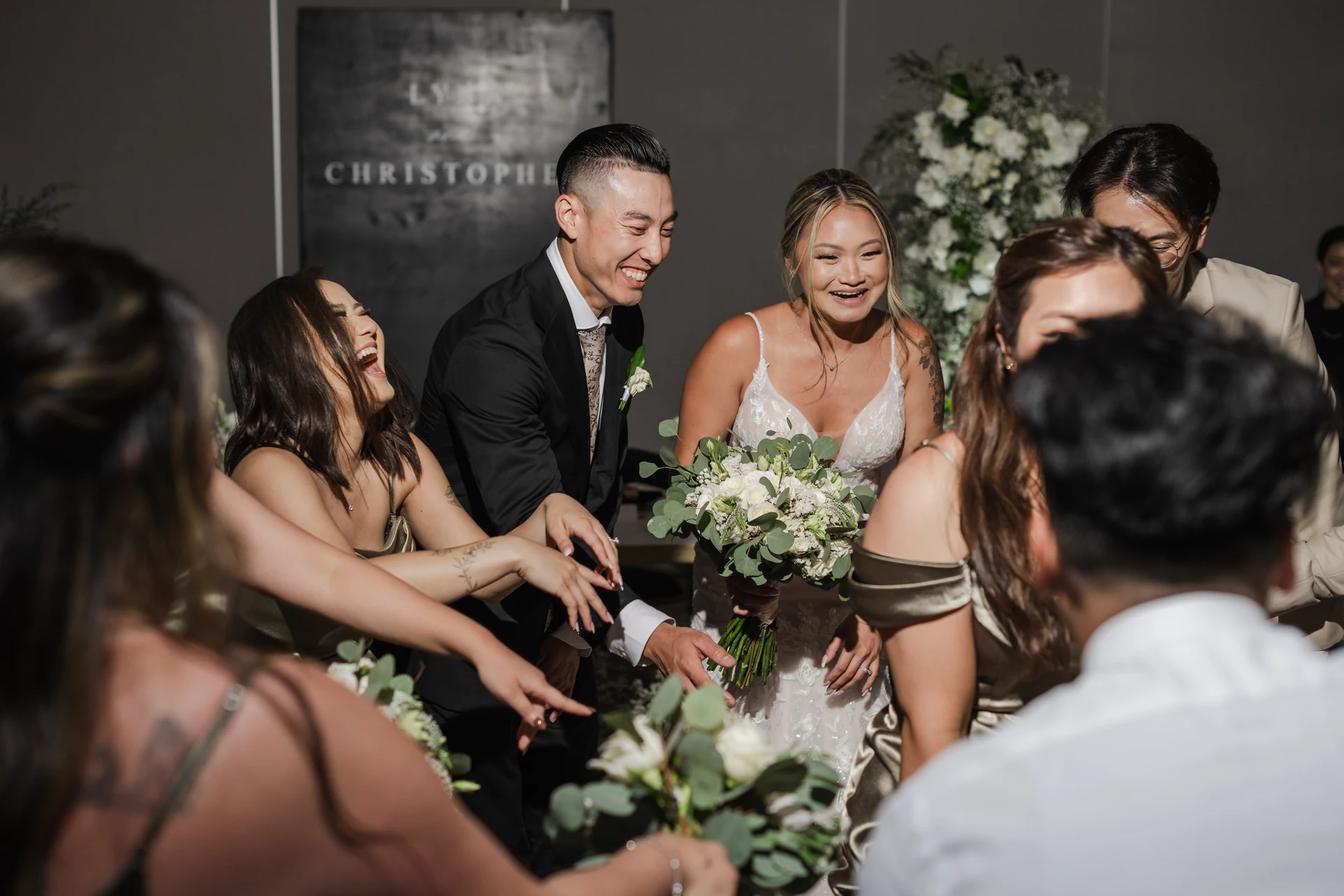Bride and groom laughing surrounded by wedding party guests reaching toward the bouquet