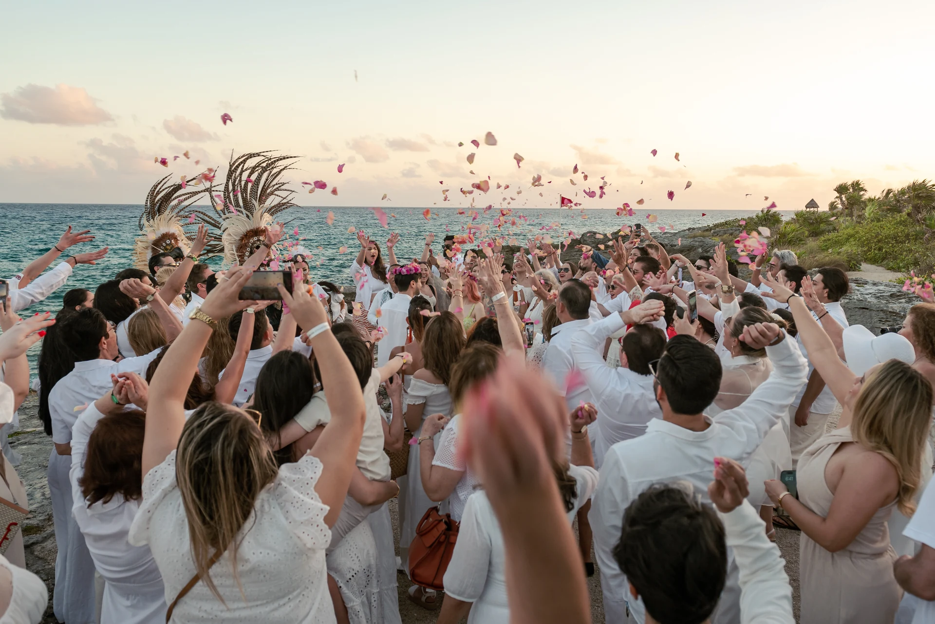 Wedding guests in white throwing rose petals into the sunset sky by the ocean alongside traditional dancers