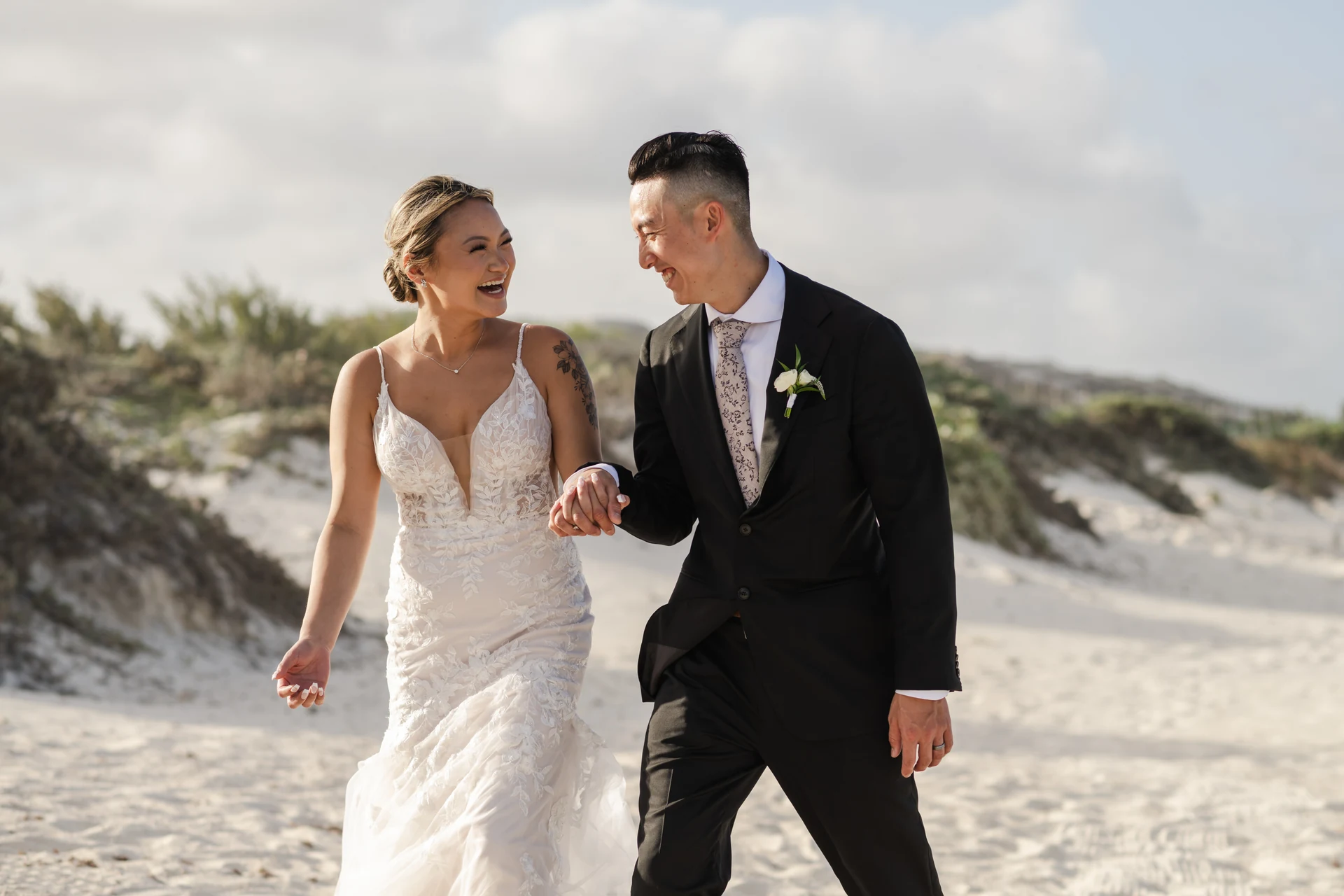 Bride and groom walking hand in hand across beach sand dunes and laughing